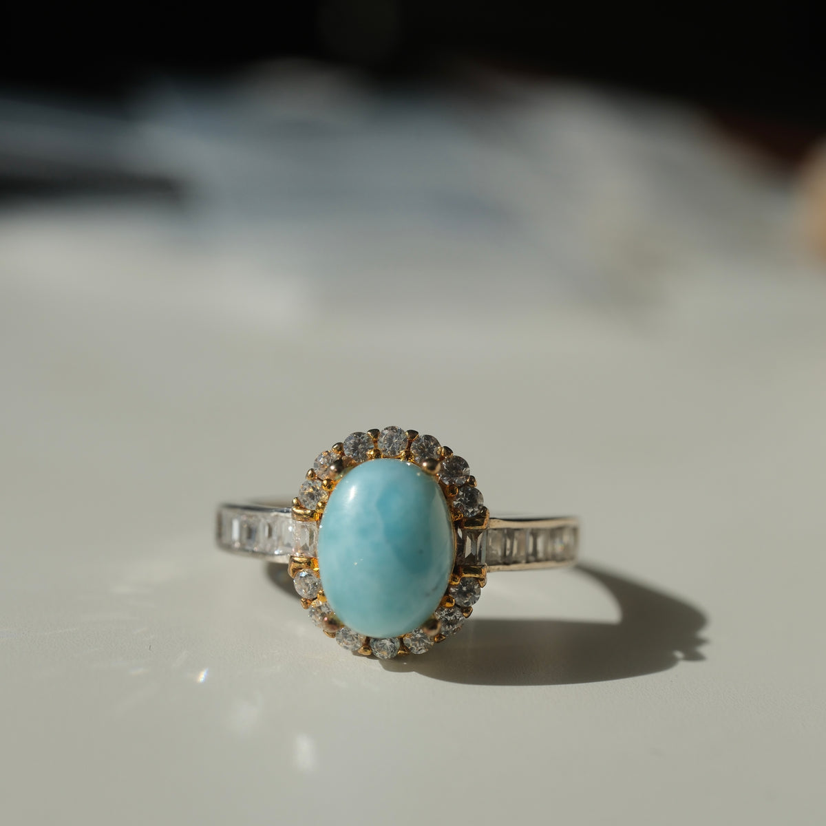 Larimar S925 adjustable ring with an oval blue stone, clear halo, gold-coloured setting details and rectangular clear shoulder accents